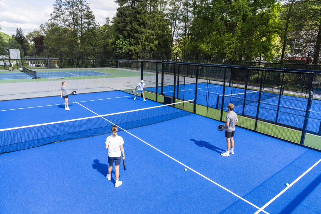 Vier Personen spielen ein Doppel-Match auf einem modernen blauen Padel-Tennisplatz im Freien, umgeben von Bäumen und Sportanlagen