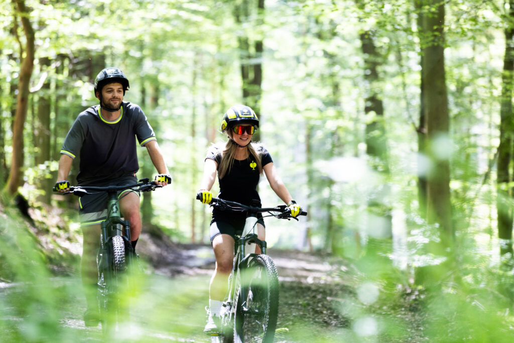 Zwei sportlich gekleidete Personen fahren mit E-Mountainbikes durch einen hellen Waldweg und genießen die Fahrt durch die Natur